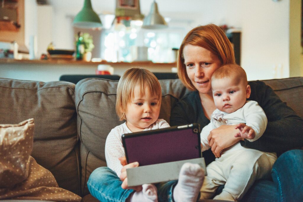 Eine Mutter sitzt mit ihrem Kleinkind und Baby auf dem Sofa und schaut sich etwas auf dem Tablet an
