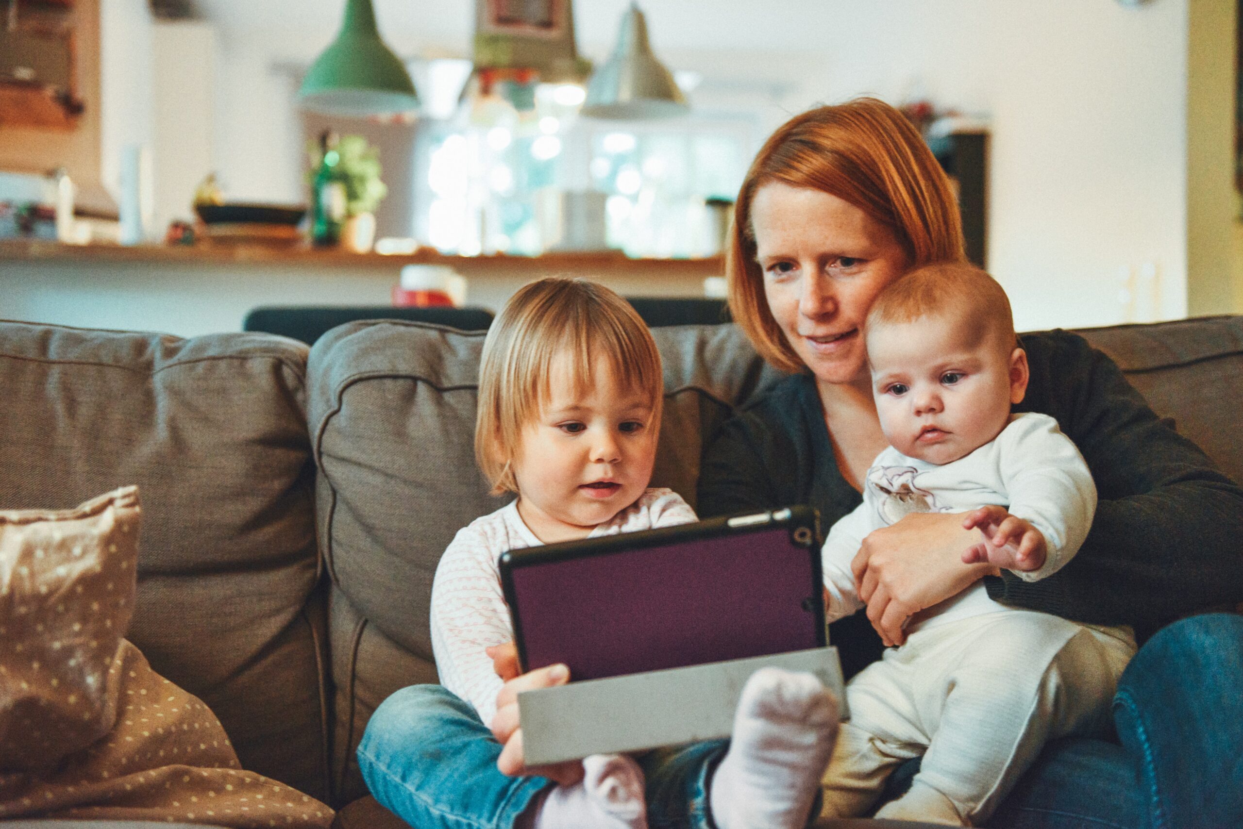 Eine Mutter sitzt mit ihrem Kleinkind und Baby auf dem Sofa und schaut sich etwas auf dem Tablet an