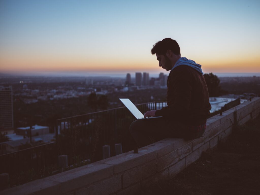 Junger Mann, der auf einer Mauer mit einem Laptop auf dem Schoß sitzt