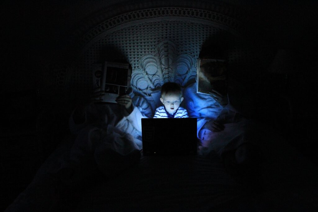 Ein kleines Kind sitzt mit einem Laptop im Bett und wird von blauem Licht angestrahlt