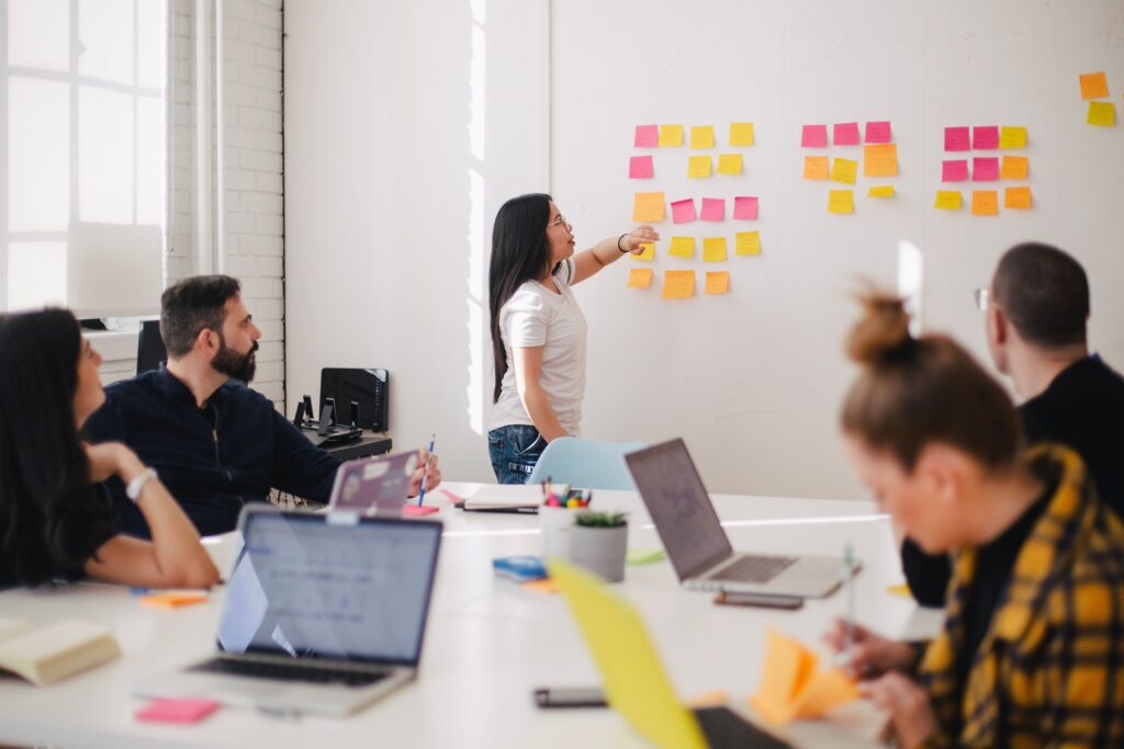 Eine junge Frau erklärt einer Gruppe etwas an einer Wand mit bunten Post-its