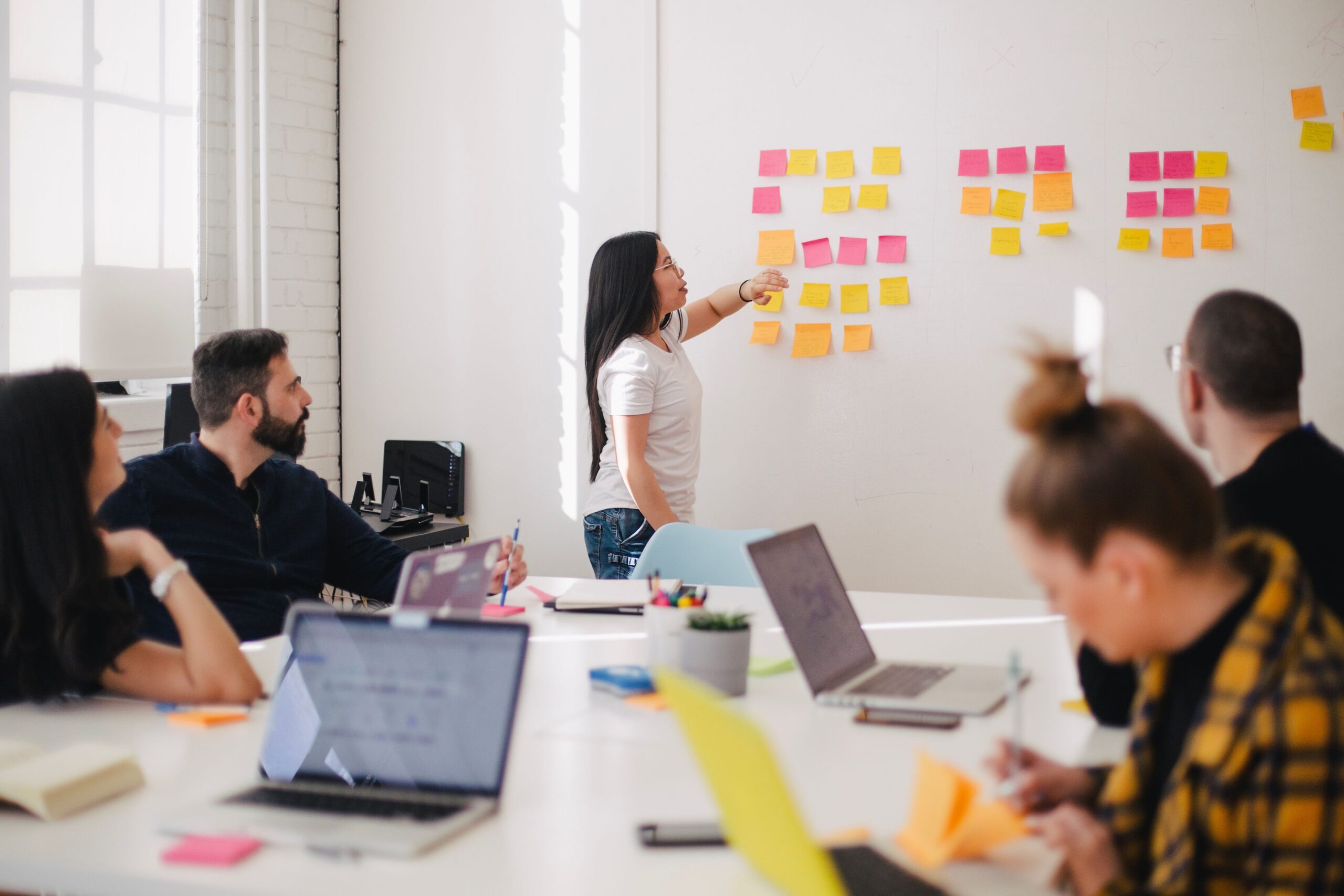 Eine junge Frau erklärt einer Gruppe etwas an einer Wand mit bunten Post-its