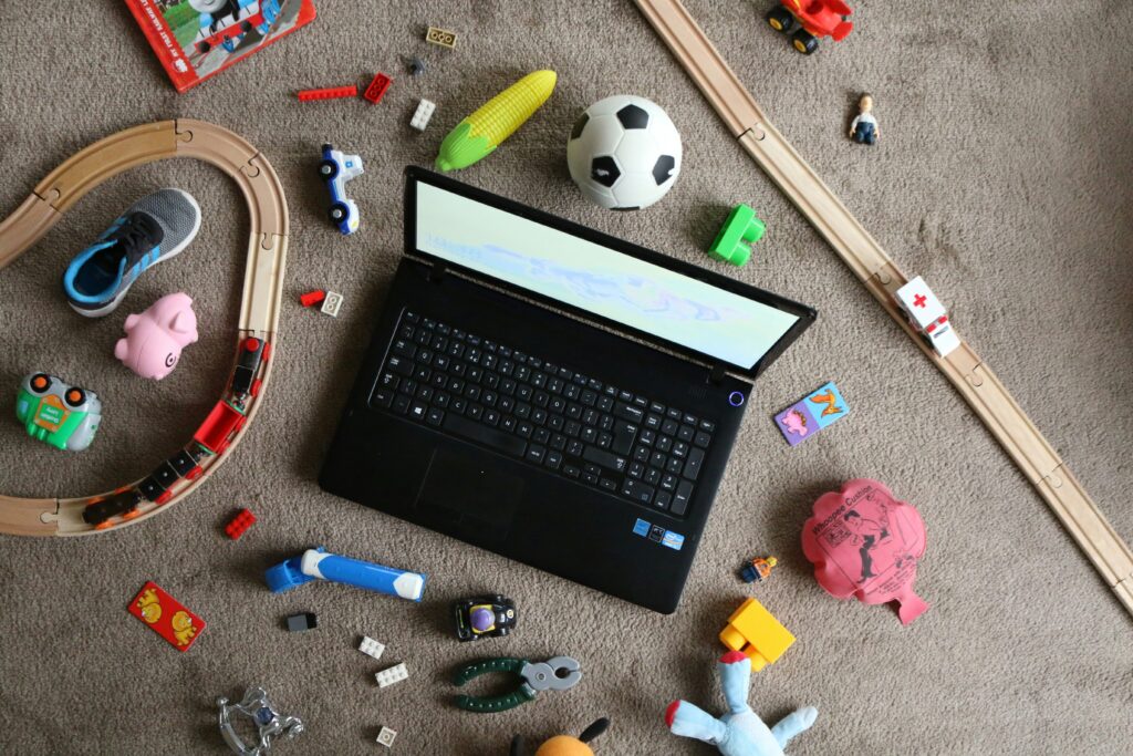 Ein aufgeklappter Laptop steht zwischen vielem Kinderspielzeug auf dem Boden.