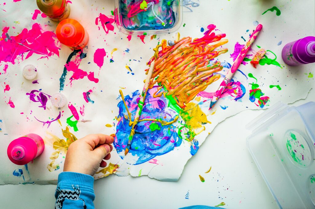 Eine Kinderhand ist vor einem bunten Bild mit Farbtuben, Pinseln und Farbklecksen zu sehen.