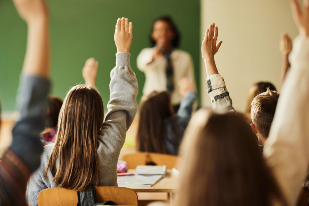 Eine Gruppe von Kindern im Klassenraum ist von hinten zu sehen. Alle heben die Hände in die Luft.