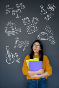 Ein junges Mädchen mit Büchern in der Hand steht vor einer schwarzen Tafel auf der verschiedene Symbole der Berufsorientierung zu sehen sind.