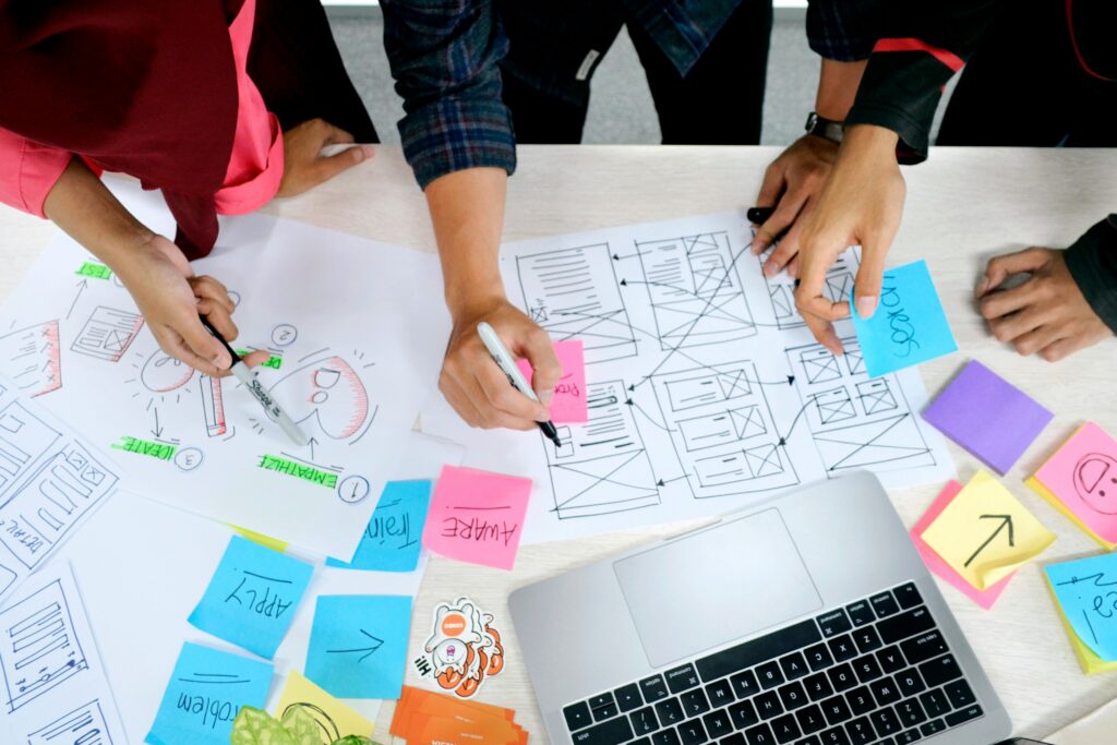 Auf einem Tisch sind ein Laptop, Schriftstücke und bunte Post-its zu sehen an denen 3 Personen arbeiten.