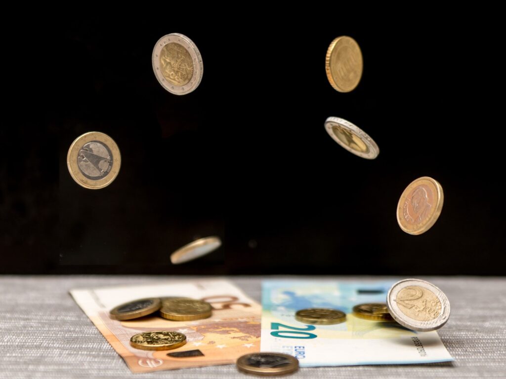 Zwei Geldscheine liegen auf einem Tisch darüber sind fliegende Münzen zu sehen.