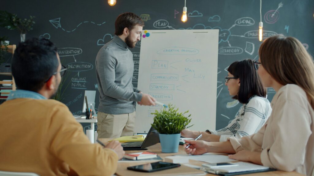 In einem Workshop erklärt eine Person an einem Flipchart 3 anderen Personen etwas.