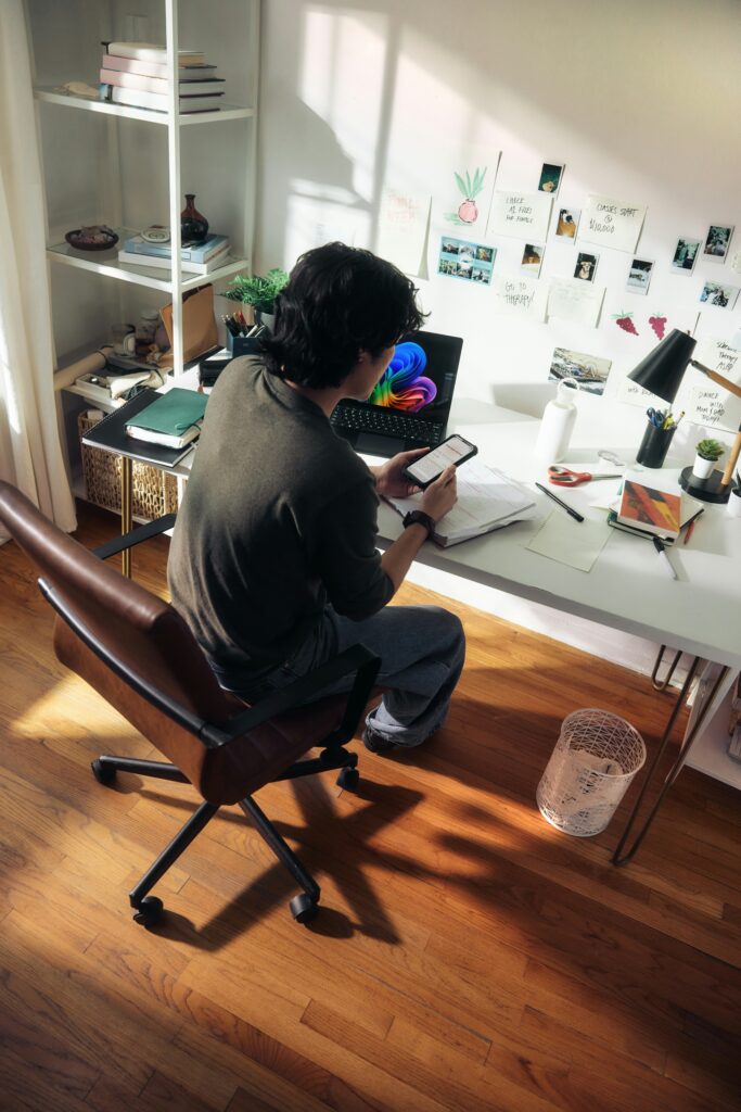 Ein junger Mann sitzt am Schreibtisch vor einem Laptop und hat ein Handy in der Hand.